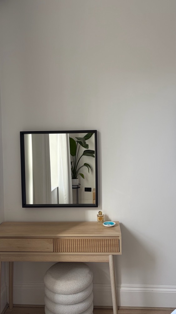 Square black mirror above a rattan dressing table and pouffe